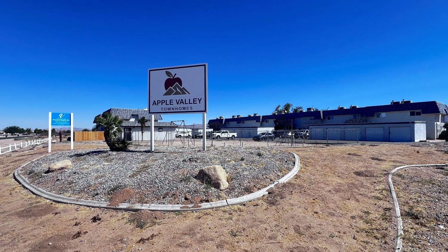 The entrance to Apple Valley Townhomes on Sioux Road: the property sign with the apple and mountain logo, the two-story townhomes and the row of garages behind it