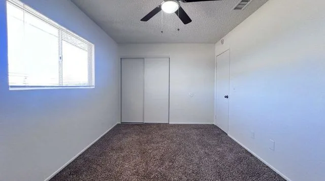 A bedroom with tan carpet, a ceiling fan, a wide window with blinds and a sliding-door closet